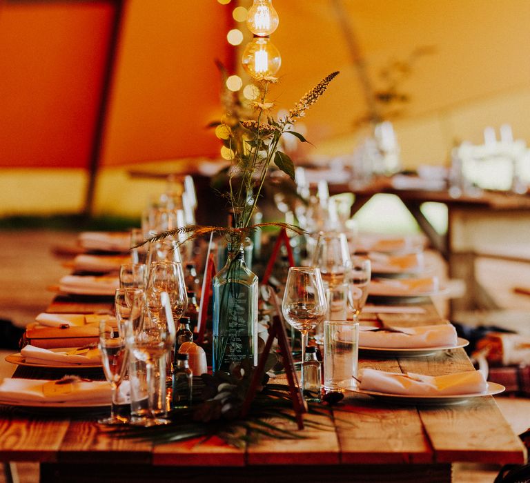 Hanging lights above wooden table complete with gin bottle vases for rustic style reception 