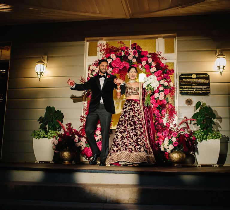 Bride & groom leave doorway covered in bright red and pink florals on their wedding day