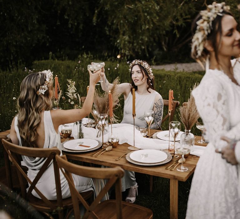 Bridesmaids in grey satin dresses and dried flower crowns drinking champagne at the intimate outdoor reception as the bride and groom dance 