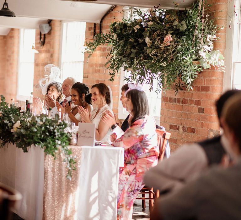 Two brides sit with wedding guests at top table under green foliage cloud with white table cloth, green foliage table runner and white candles in red brick industrial wedding reception room at The West Mill Derby