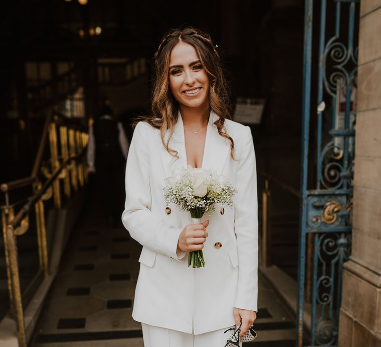 Bride in a white trouser suit and blazer holding a white rose and gypsophila bouquet 