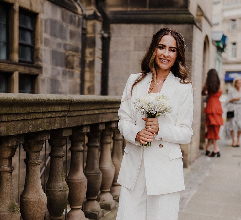 Stylish bride in a three-piece white suit with jacket and wide leg trousers holding a white rose bouquet 