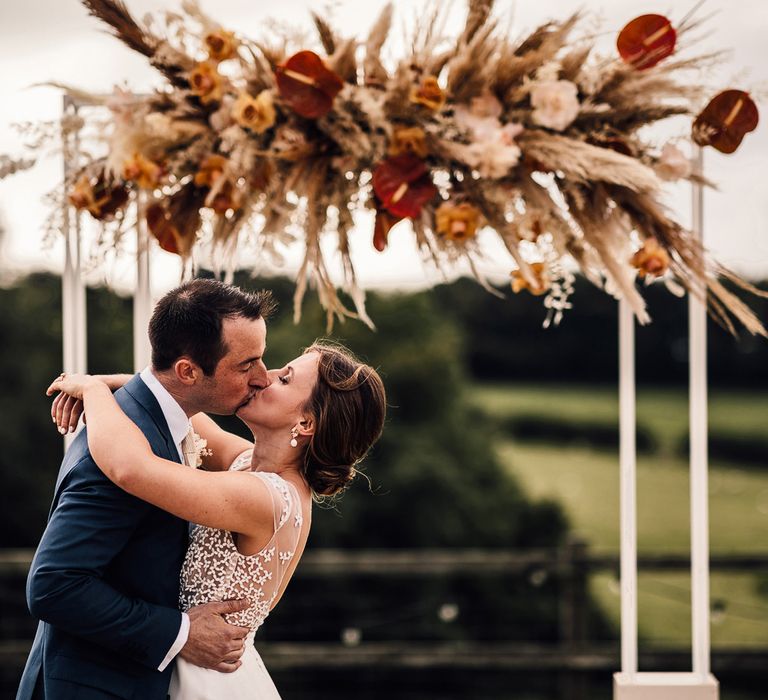 Groom in blue suit kisses bride with wedding updo wearing white Rime Arodaky wedding dress in front of white rectangular wedding arch with pampas grass flower cloud during outdoor wedding ceremony in Dorset