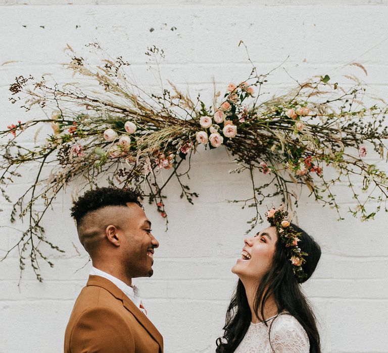 Bride in a long sleeve lace wedding dress and flower crown laughing with her groom in a light brown coloured suit under a twig, dried grasses and pink flower installation