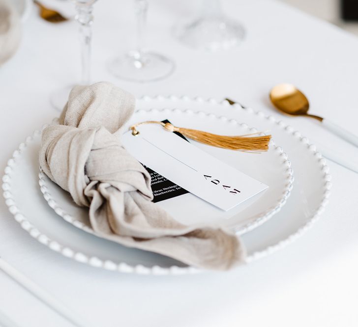 A black and white wedding place setting has two plates, a neutral coloured tied napkin, gold cutlery, place tag with gold tassel and tall white tapered candles.