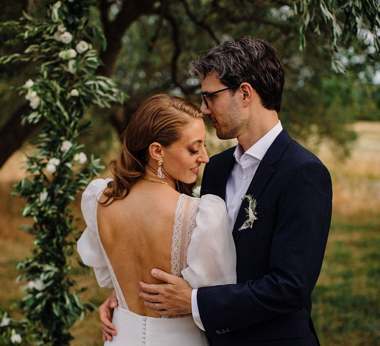 Bride wears Marta Marti wedding gown with lace trip low-cut back as groom places his hand behind her whilst wearing suit with open shirt