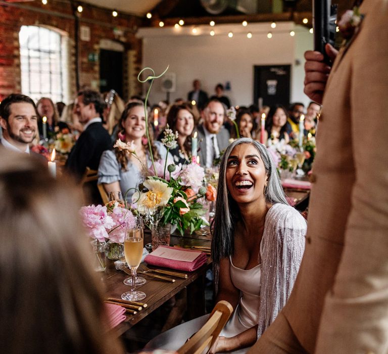 Bride in satin Halfpenny London wedding dress and tasselled cape smiles up at groom in double breasted linen suit as he makes speech during wedding breakfast at Loft Studios London