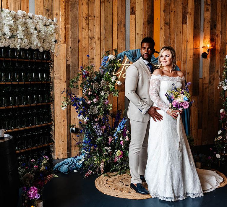 Portrait of the groom in beige suit and bride in a lace Bardot wedding dress standing next to their sparkling wine wall 