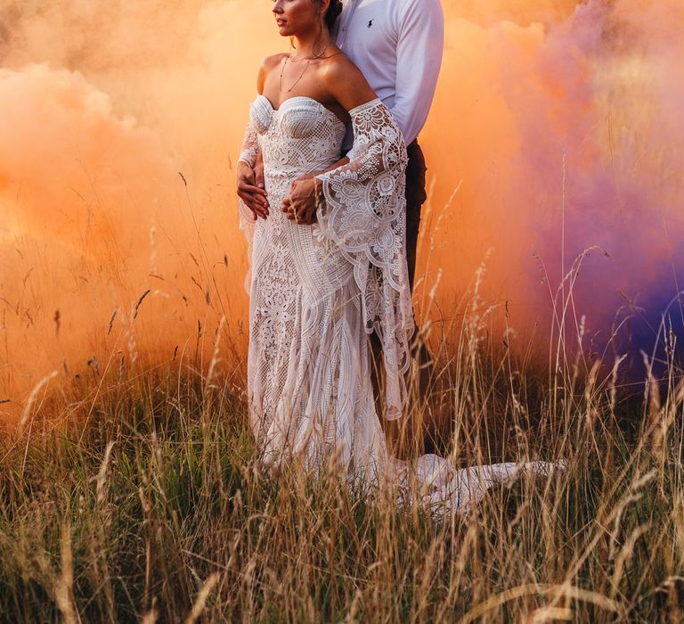 Groom in an open collar white shirt embracing his boho bride in a strapless lace wedding dress with an orange smoke bomb going off in the background