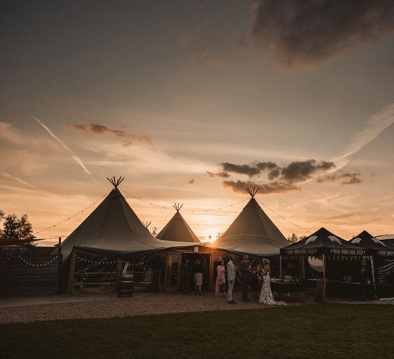 Wedding guests stand outside of tipi during sunset at Inkersall Grange Farm wedding