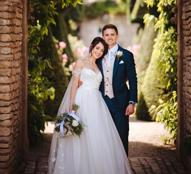 Bride leans her head into groom on their wedding day as they stand beneath archway surrounded by green foliage