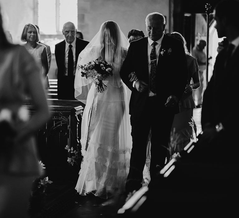 Bride in Halfpenny London Mayfair dress and veil holding mixed bridal bouquet walks down the aisle arm in arm with father in suit during church ceremony in Cornwall