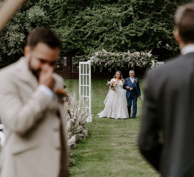 Bride in homemade wedding dress holding rose bridal bouquet walks up the aisle holding arm of man in blue three piece suit during DIY garden wedding ceremony in Bedfordshire