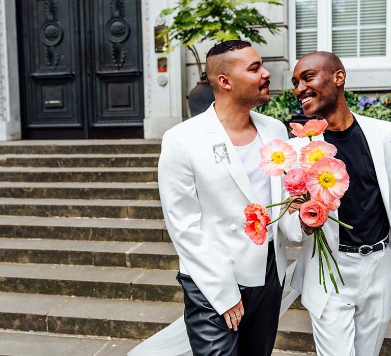 Stylish grooms in white satin jackets holding a red anemone bouquet at their city elopement 