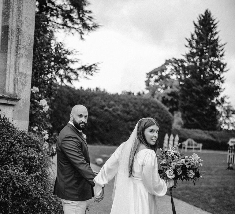 Black & white image of bride & groom walk hand in hand with one another on their wedding day