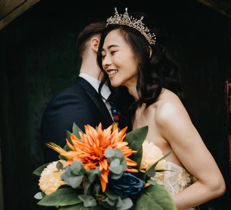 Smiling bride in headpiece and strapless wedding dress holds tropical wedding bouquet as groom in dark suit hugs her