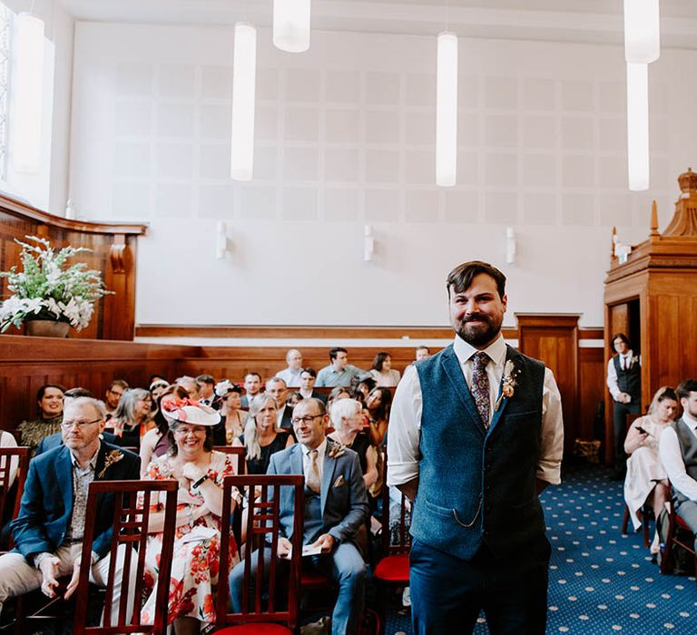 Groom waits for his bride at the end of the aisle and wears blue waistcoat with white shirt