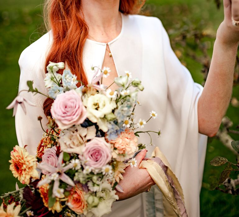 Bride in white wedding dress and cape holds multicoloured bridal bouquet wearing white headband with net holds apple as she stands in orchard