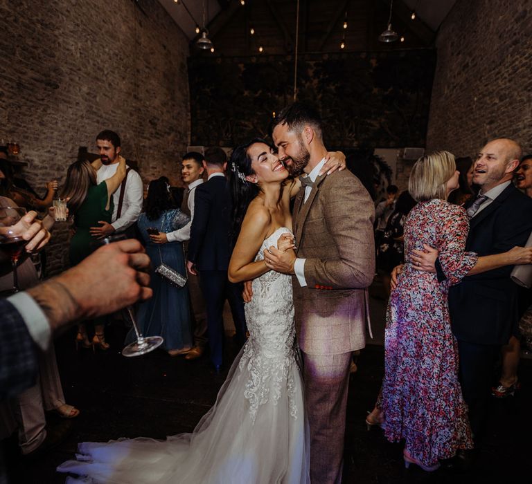 Bride in lace and tulle wedding dress with train dances with groom in brown tweed suit during stone barn wedding reception in the Costwolds