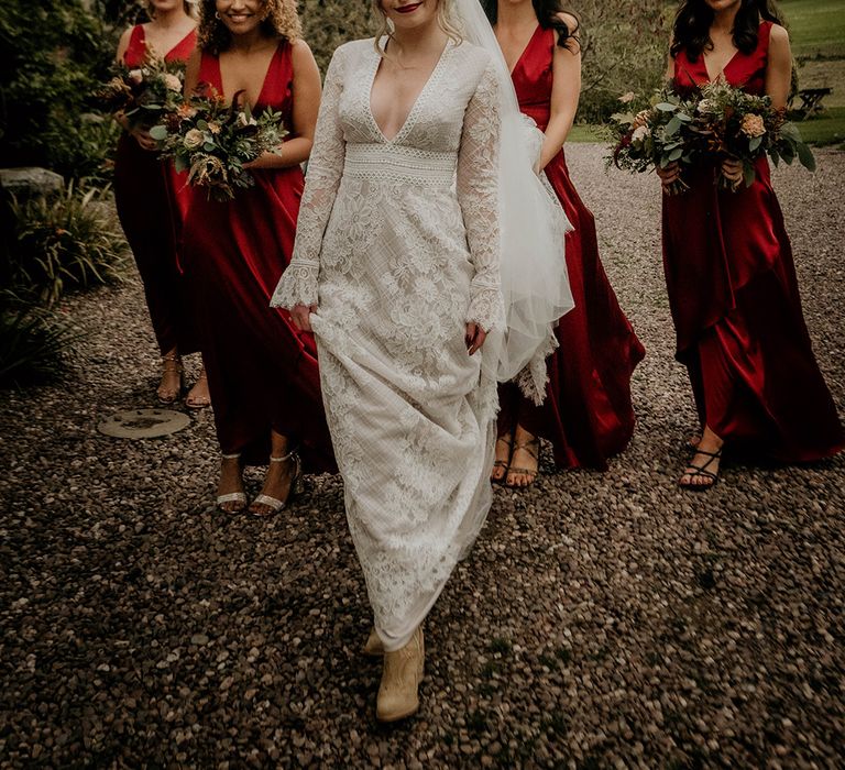 Boho bride in a lace wedding dress with plunging neckline and long lace bell sleeves standing with her bridesmaids in satin red dresses 