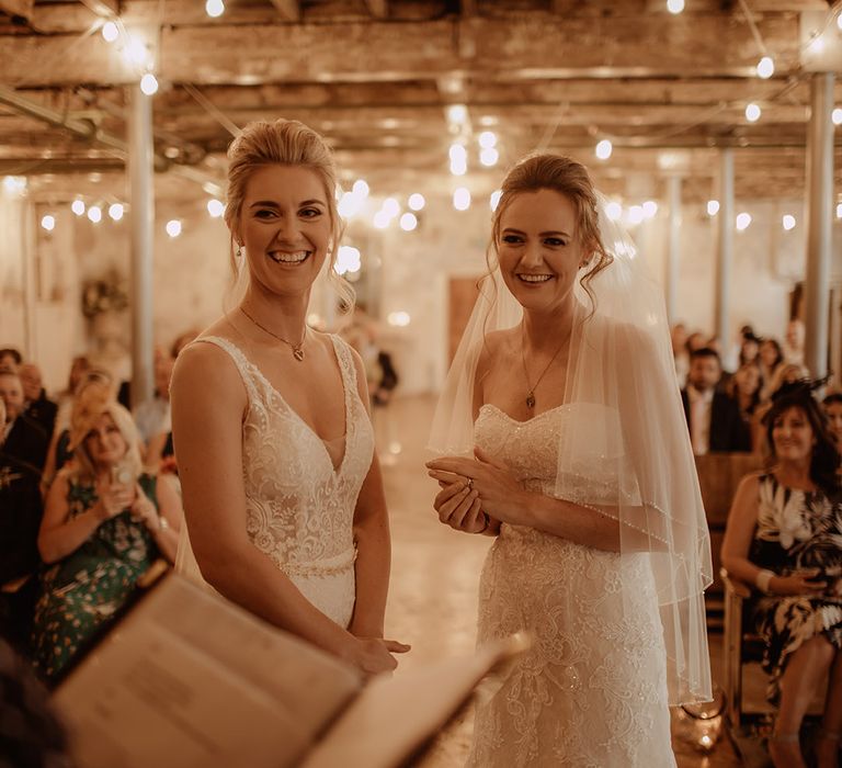 Brides at the altar on their wedding day as fairy lights hang from the ceiling behind them