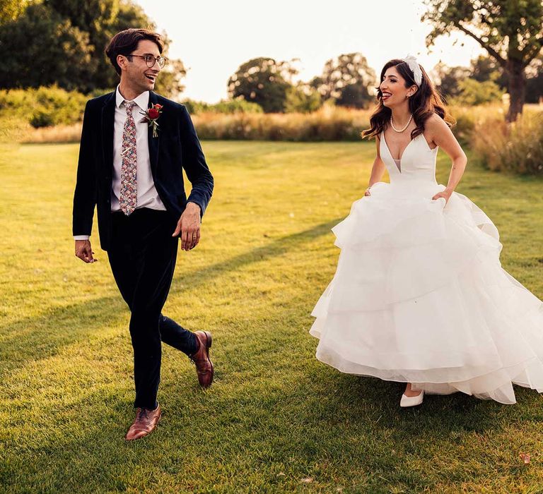 Bride in princess wedding dress with layered tulle skirt and feathered headband walks through grounds with groom in dark blue suit and floral tie during golden hour