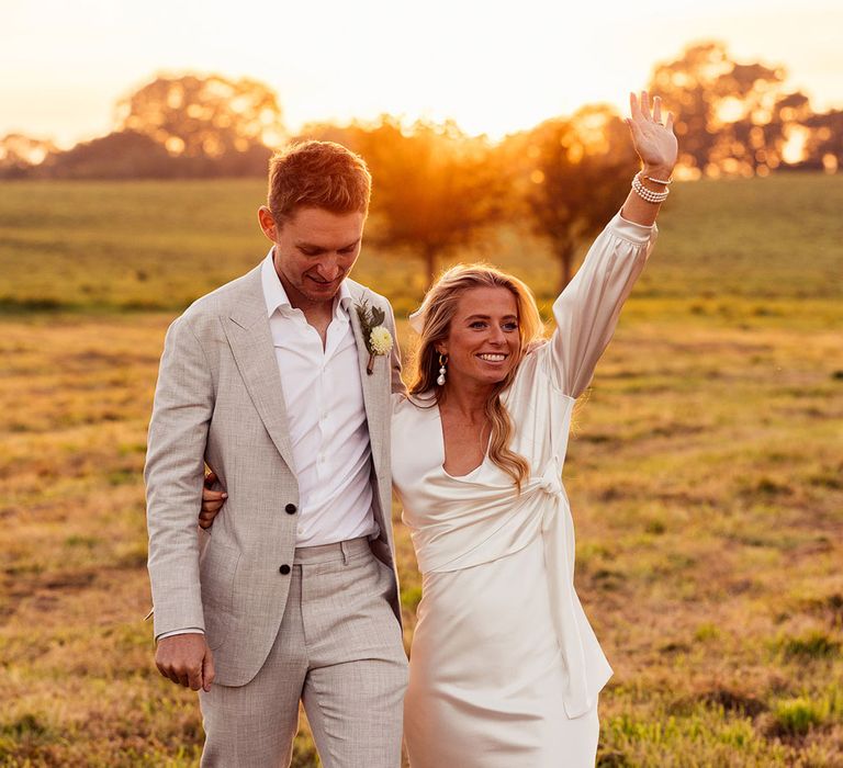 Bride in white satin wedding dress and wrap top throws arm in the air as she walks with groom in light grey suit and white open shirt 