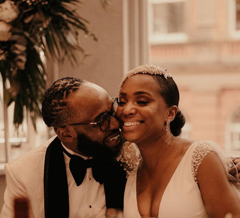 Groom in a white tuxedo jacket kissing his bride in a beaded cap headdress at their vintage style city wedding reception