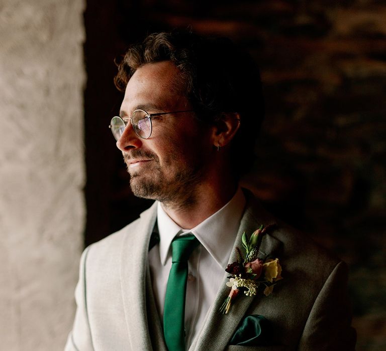 Groom in bespoke light grey suit with deep green tie and handkerchief and colourful flower boutonnière with round glasses