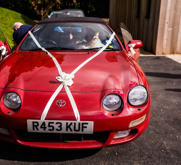 Bride in bright red vintage Toyota wedding car transportation with white ribbon