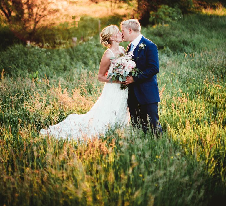 Groom in blue suit and light blue tie kisses bride in boho lace wedding dress and pink flower wedding bouquet 