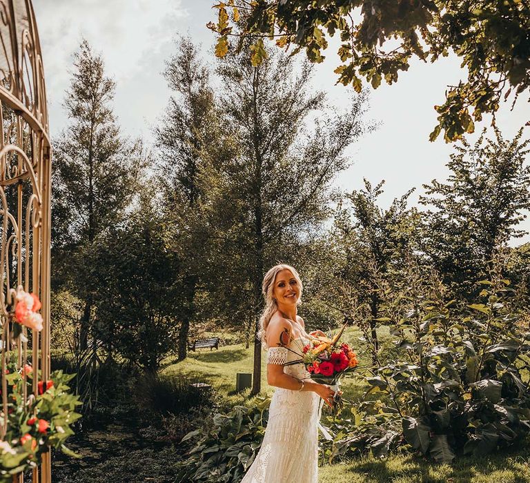 Bride in strapless lace boho wedding dress with train and holding red and orange flower bouquet