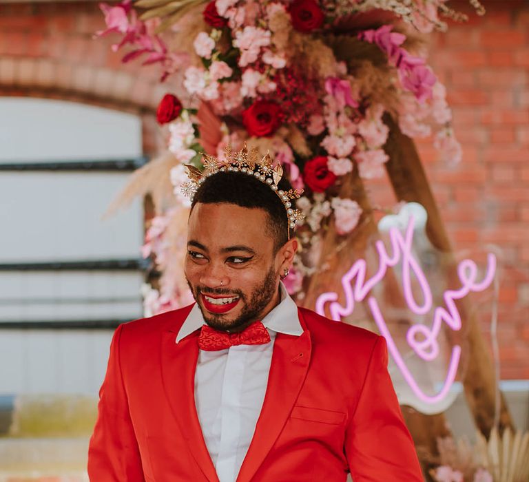 Groom in a red suit and bow tie wearing black winged eyeliner and a gold crown accessory 