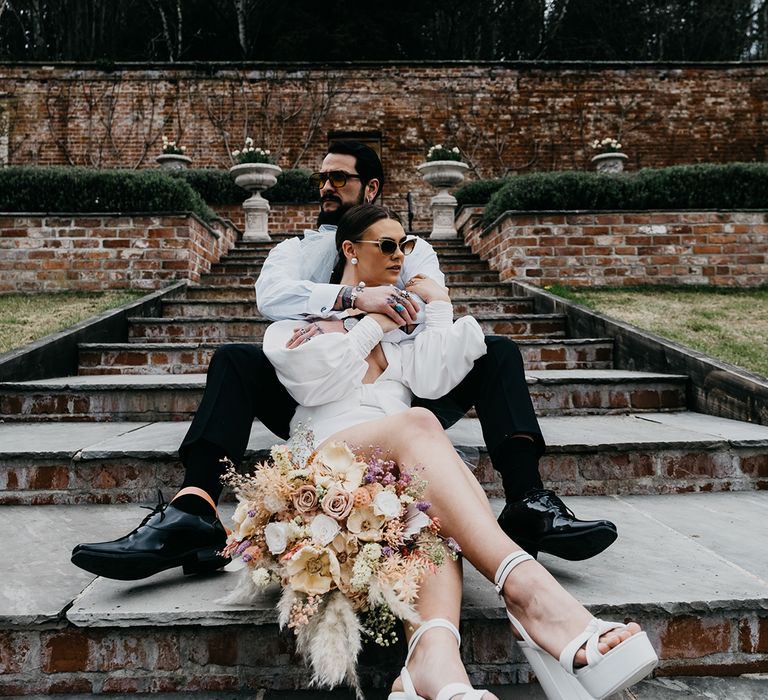 Groom with rings and tattoos embracing his bride in a short wedding dress with platform shoes and sunglasses next to her preserved pastel flowers and dried grasses wedding bouquet 