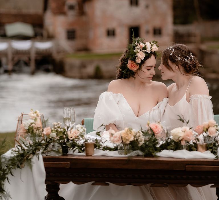 Two brides in ethereal wedding dresses sitting at their utdoor wedding reception table at Mapledurham with white drapes and a foliage and pink flower table runner 