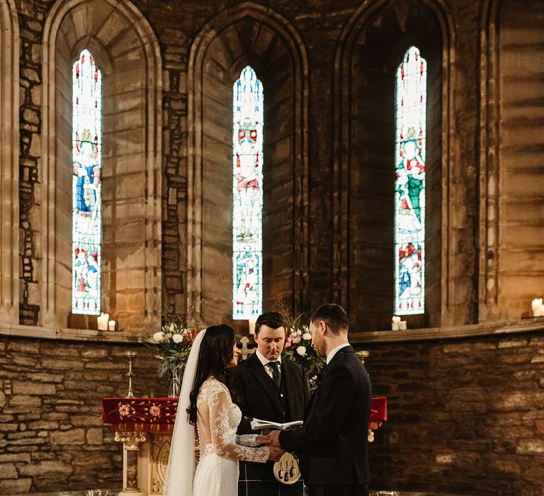 Bride wears floor length veil and holds the hands of her groom in front of stained glass windows during church ceremony 