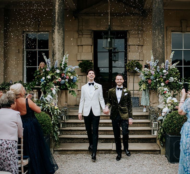 Grooms hold hands after wedding ceremony and walk beneath confetti falling above them 