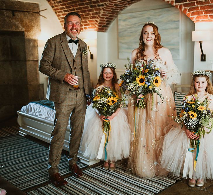 Bride wearing pink starry wedding dress stands beside flower girls who wear light pink dresses with ballerina skirts and floral crowns holding sunflower bouquets