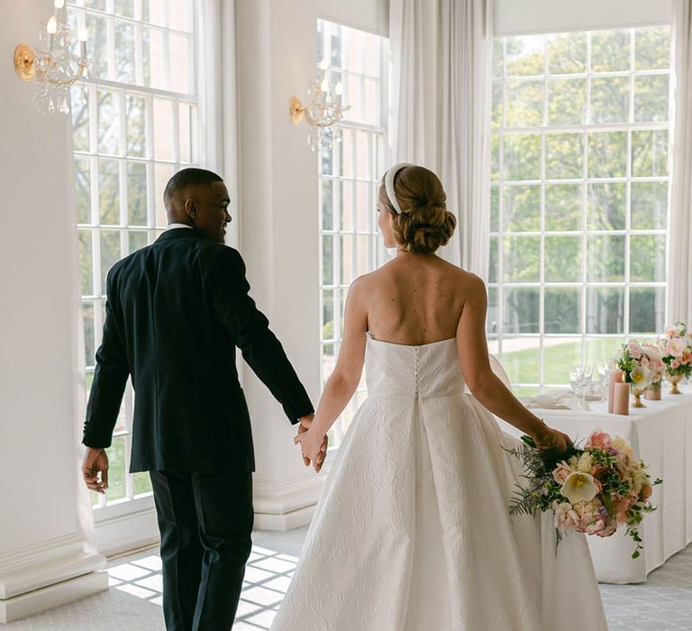 Bride wears a beaded headband with her updo hairstyle with a strapless brocade wedding dress with groom in black tie walk into their wedding breakfast