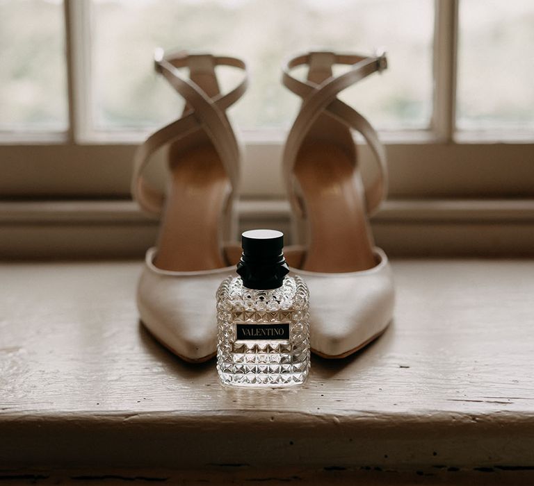 Valentino perfume in front of satin white pointed heeled bridal shoes with crossover straps to the front