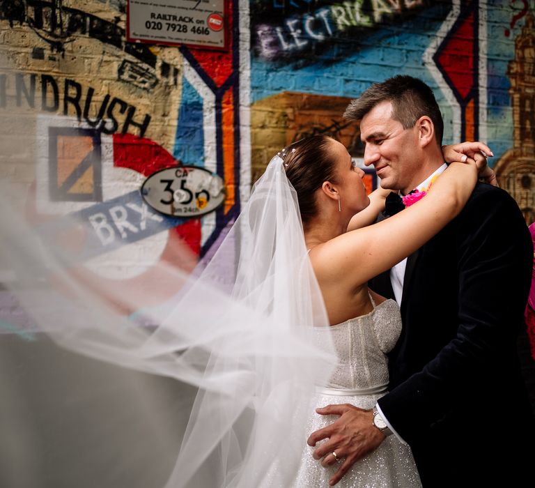 Brides veil blows in the wind as she stands with her groom in front of colourful graffiti wall in Brixton 