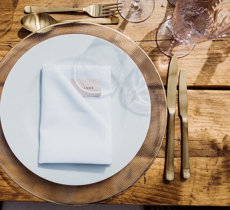 Gold cutlery with gold chargers, white plates and pink glassware and name place cards for the table settings 