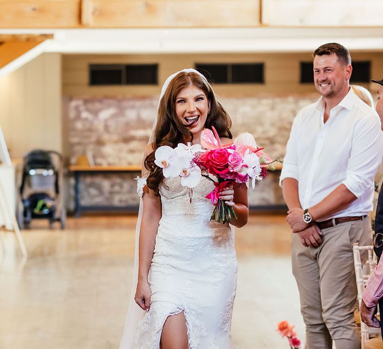 Bride walks down the aisle wearing floor-length veil and off-the-shoulder lace wedding dress whilst holding bright pink floral bouquet 