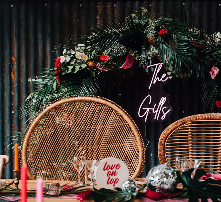 Mismatched retro peacock chairs with a custom pink neon sign and tropical flowers and palm leaves