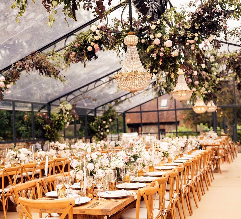 Glass marquee with chandelier installations with pink and white rose flowers, and long wooden banquet tables 