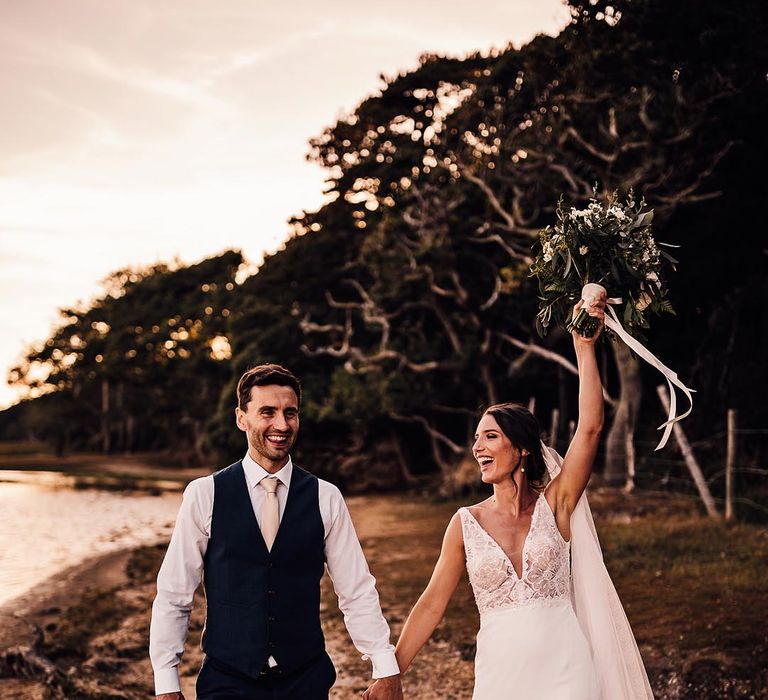 The groom in a navy waistcoat walks holding hands with the bride in a Blue by Enzoani wedding dress