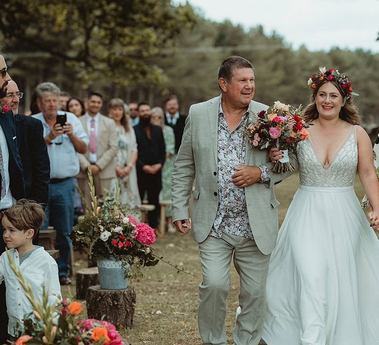 Bride wearing a flower crown and holding a colourful bouquet is walked down the aisle by her mother and father in light green outfits 