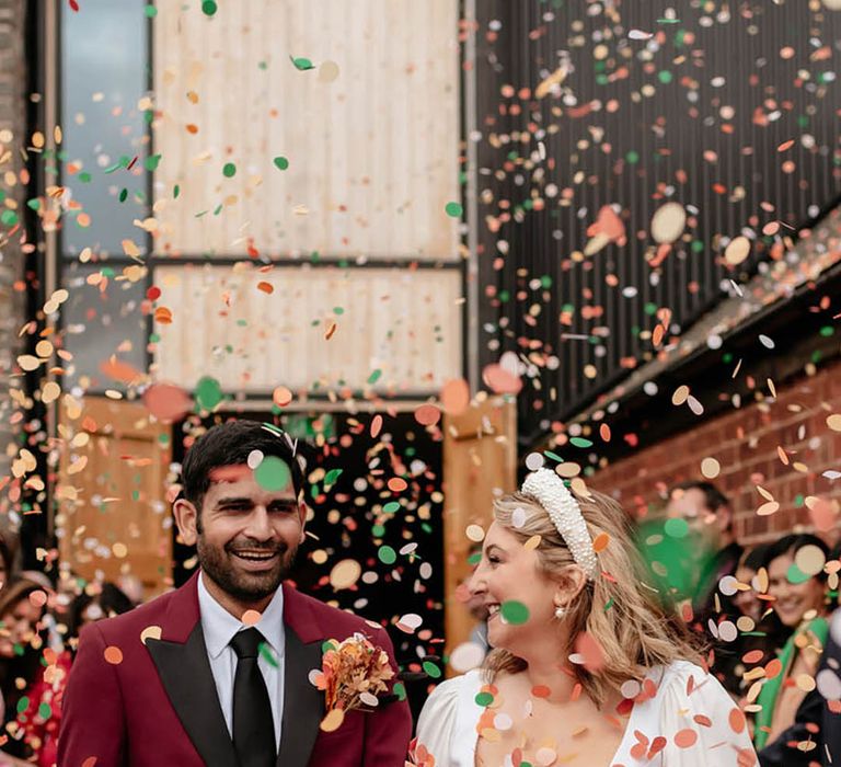 Smiling groom in burgundy suit with black tie and black lapel detail walking with the bride in a square neckline wedding dress with confetti 