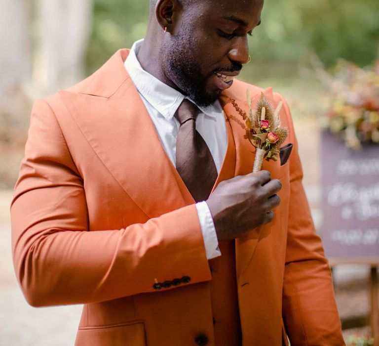 Groom adjusting his dried flower, rose and pampas grass boutonniere wearing burnt orange rust coloured suit with brown tie, pocket square and brown accessories 