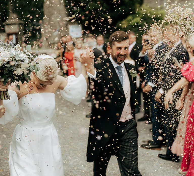 Bride & groom in morning suit and pale blue tie walk through colourful confetti exit 